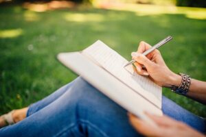 an image of a person sat down on green grass writing how to plan the best camping trip in a notebook